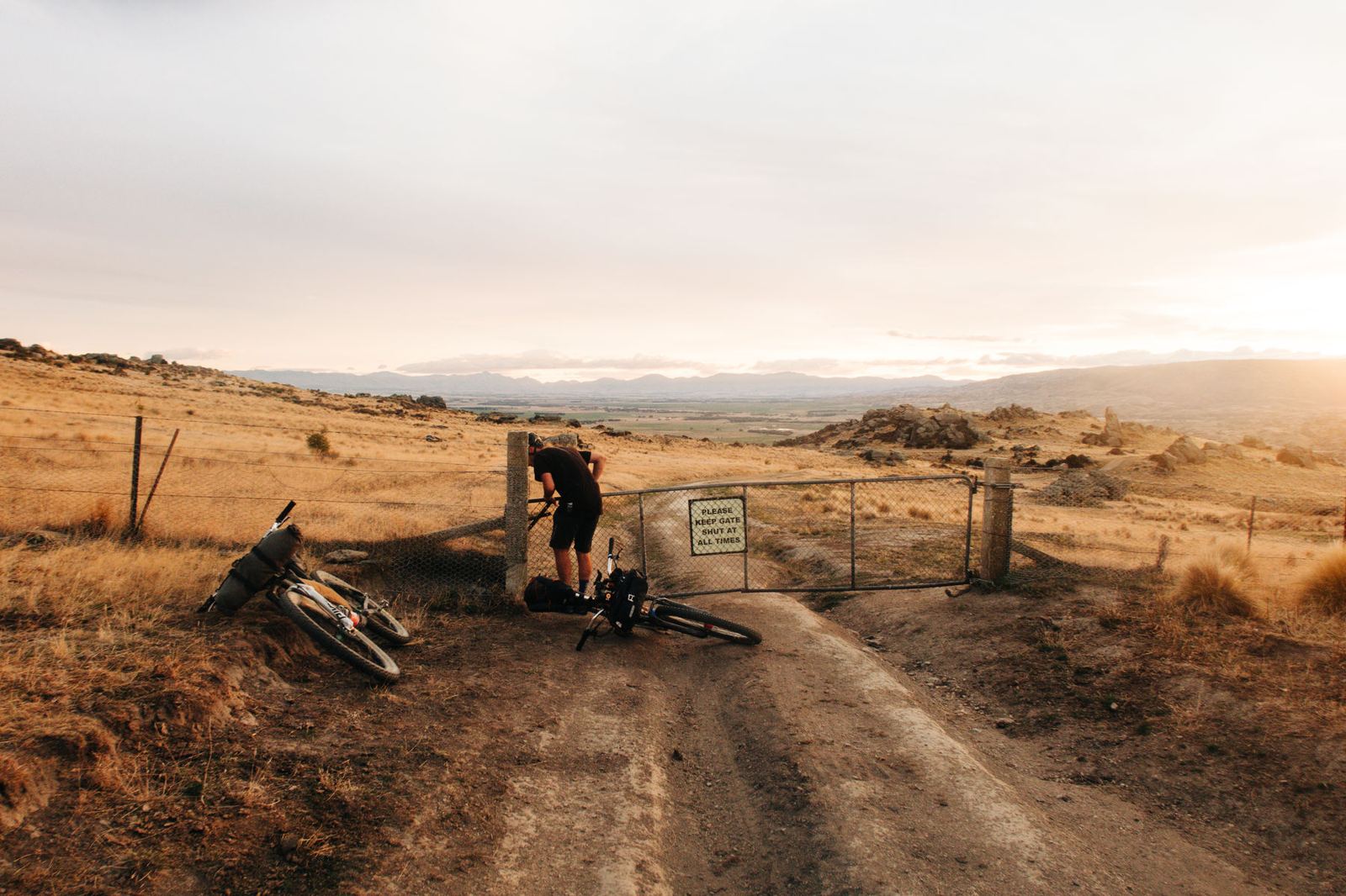 Two riders stopped at a farm gate on a high-country track at dusk